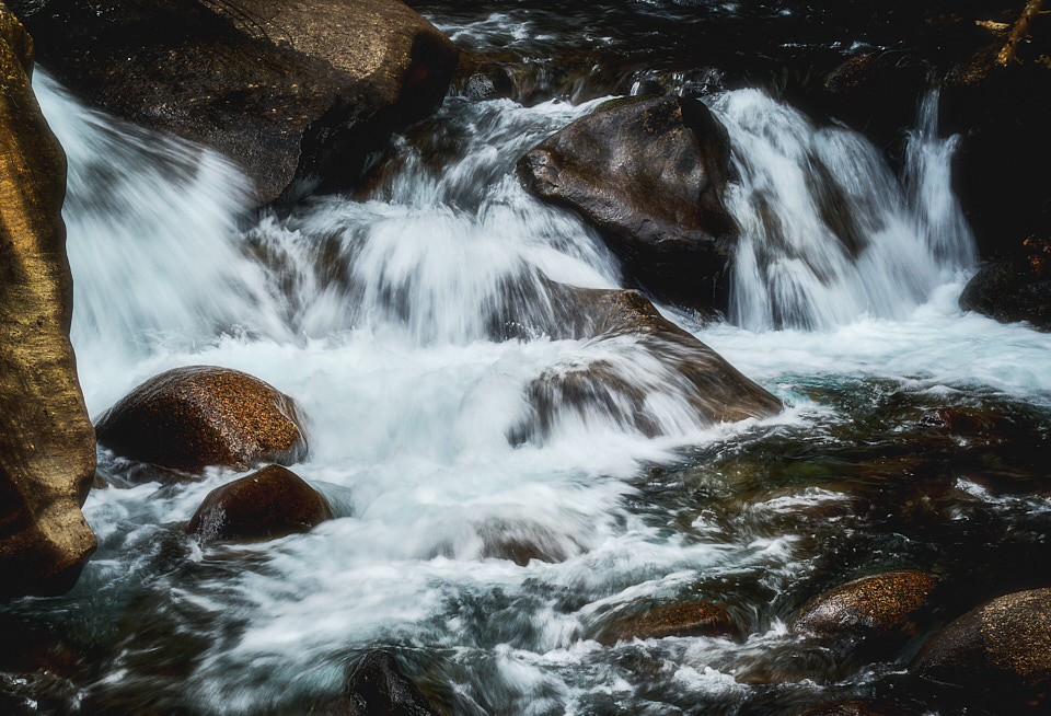 Clear mountain water rushes over and around dark, smooth boulders, forming small cascades and white foam.